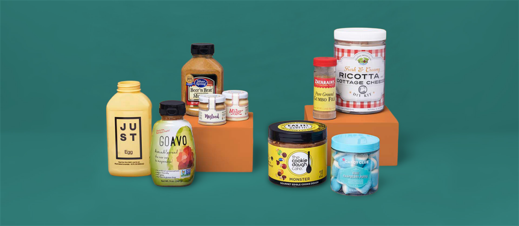 Assorted gourmet food bottles and jars arranged on orange display blocks against a teal background (Mustard bottle, egg product, sauces, cookies, and sci-enth jars).