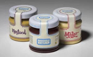 Three small glass jars of condiments (ketchup, mustard, and mayonnaise) with white lids and vintage-style labels featuring checkered tape.