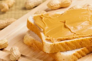Slices of bread with creamy peanut butter spread on top, placed on a wooden cutting board with peanuts around it.