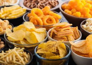 Assorted bowls of crunchy snacks, including potato chips, tortilla chips, pretzels, popcorn, and cheese puffs.