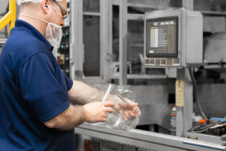 Factory worker in blue uniform and hairnet handling glass jars on a production line with industrial machinery and a monitor nearby