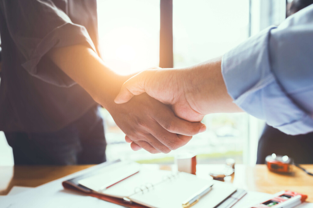 Two people shake hands over a desk in a bright office, with papers and a phone nearby—symbol of agreement or collaboration.