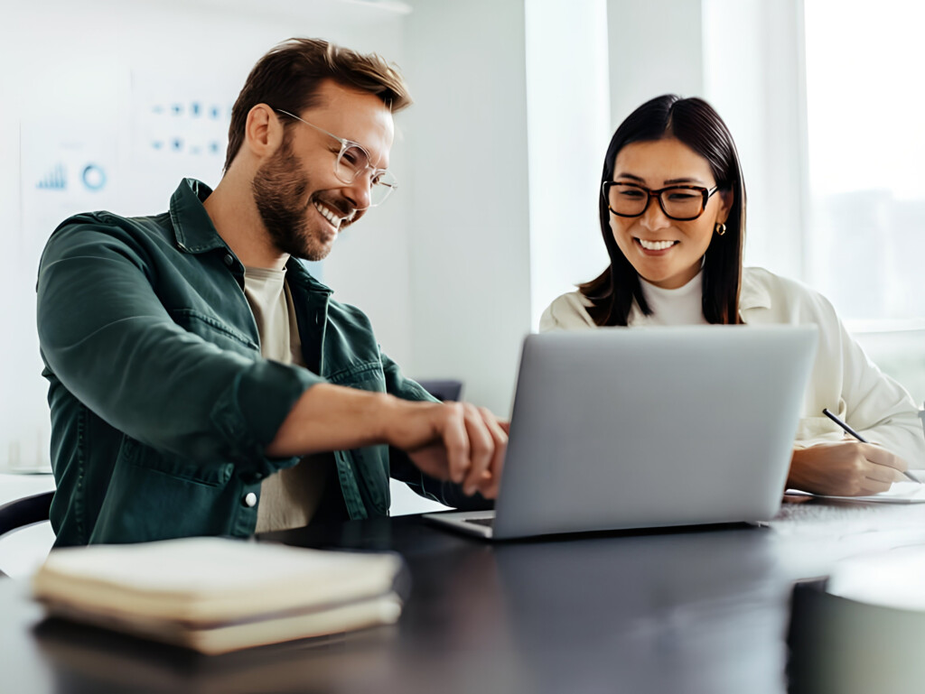 Two coworkers smiling while looking at a laptop at a bright office desk.